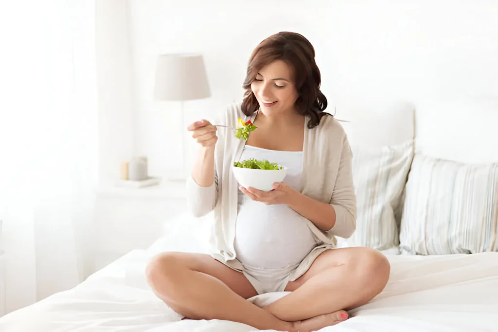 Pregnant person in a sunny kitchen holding a water bottle and a bowl of fresh salad, smiling warmly.